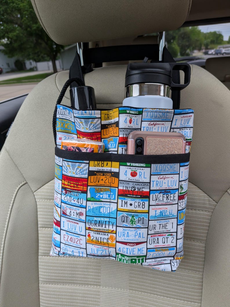 K&ouml;nnte beinhalten: Autositz-Organizer mit farbenfrohem Nummernschildmuster. Enth&auml;lt mehrere F&auml;cher mit einer schwarzen Wasserflasche, einem Edelstahlbecher, einem Telefon und anderen Gegenst&auml;nden. Der Organizer wird an der Kopfst&uuml;tze aufgeh&auml;ngt.