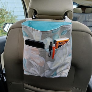 K&ouml;nnte beinhalten: Ein Autositz-Organizer mit einem floralen Muster in Grau, Wei&szlig; und T&uuml;rkis. Der Innenraum ist t&uuml;rkis. Der Organizer hat Taschen mit einem Telefon, Stiften und einem Taschentuchpaket. Er h&auml;ngt an der Kopfst&uuml;tze.