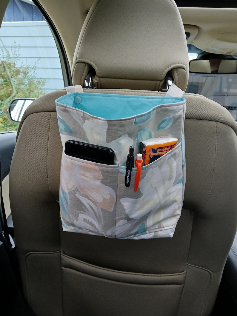 K&ouml;nnte beinhalten: Ein Autositz-Organizer mit einem floralen Muster in Grau, Wei&szlig; und T&uuml;rkis. Der Innenraum ist t&uuml;rkis. Der Organizer hat Taschen mit einem Telefon, Stiften und einem Taschentuchpaket. Er h&auml;ngt an der Kopfst&uuml;tze.