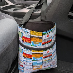 K&ouml;nnte beinhalten: Eine Auto-M&uuml;lltasche mit schwarzem Riemen und Innenraum. Die Au&szlig;enseite ist mit farbenfrohen Nummernschildern aus verschiedenen Bundesstaaten gemustert. Die Tasche befindet sich in einem Auto zwischen den Sitzen.