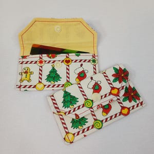 May include: Two rectangular fabric pouches featuring a white background with a repeating Christmas pattern of gingerbread people, green pine trees, red stockings, poinsettias, and candy cane stripes. Each pouch has a snap closure and yellow lining.