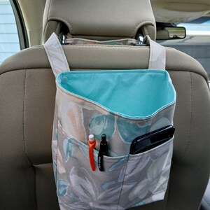 K&ouml;nnte beinhalten: Ein Autositz-Organizer mit einem floralen Muster in Grau-, Blau- und Pfirsicht&ouml;nen. Der Organizer hat eine t&uuml;rkisblaue Innenseite und mehrere Taschen mit Stiften und einem Smartphone. Er h&auml;ngt an der Kopfst&uuml;tze.