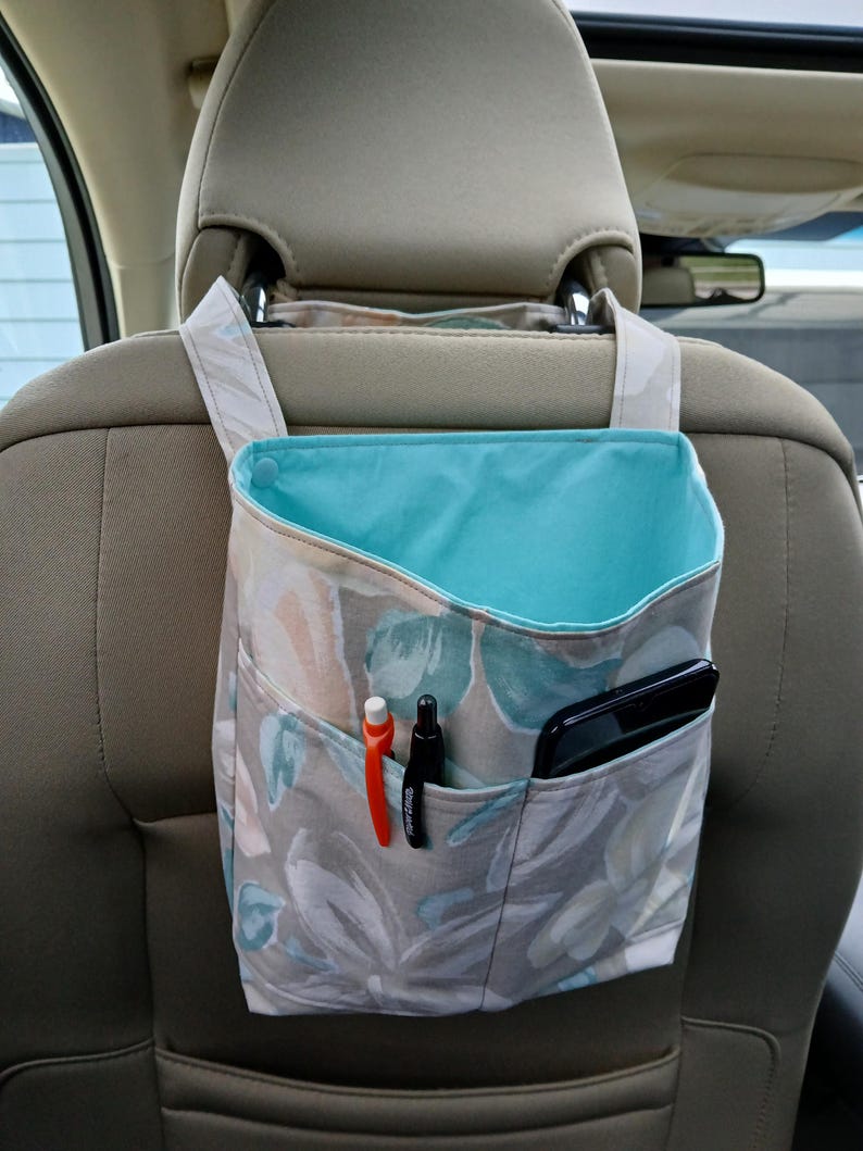 K&ouml;nnte beinhalten: Ein Autositz-Organizer mit einem floralen Muster in Grau-, Blau- und Pfirsicht&ouml;nen. Der Organizer hat eine t&uuml;rkisblaue Innenseite und mehrere Taschen mit Stiften und einem Smartphone. Er h&auml;ngt an der Kopfst&uuml;tze.
