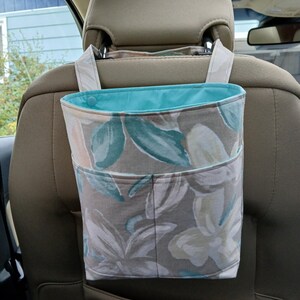 K&ouml;nnte beinhalten: Eine Autotasche mit floralem Muster und t&uuml;rkisfarbenem Innenfutter. Die Tasche besteht aus Stoff mit einem Design aus wei&szlig;en, t&uuml;rkisfarbenen und pfirsichfarbenen Blumen und Bl&auml;ttern auf grauem Hintergrund. Sie h&auml;ngt an der Kopfst&uuml;tze eines Autositzes.