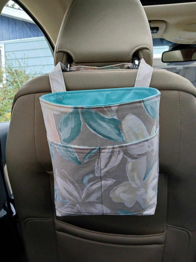 K&ouml;nnte beinhalten: Eine Autotasche mit floralem Muster und t&uuml;rkisfarbenem Innenfutter. Die Tasche besteht aus Stoff mit einem Design aus wei&szlig;en, t&uuml;rkisfarbenen und pfirsichfarbenen Blumen und Bl&auml;ttern auf grauem Hintergrund. Sie h&auml;ngt an der Kopfst&uuml;tze eines Autositzes.