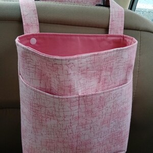 K&ouml;nnte beinhalten: Rosafarbener Auto-Organizer mit strukturiertem Muster. Der Organizer hat eine gro&szlig;e Tasche und eine kleinere Tasche auf der Vorderseite. Er h&auml;ngt an der Kopfst&uuml;tze eines Autositzes.