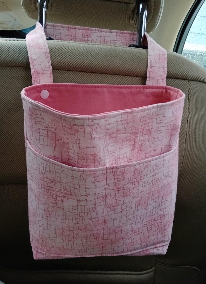 K&ouml;nnte beinhalten: Rosafarbener Auto-Organizer mit strukturiertem Muster. Der Organizer hat eine gro&szlig;e Tasche und eine kleinere Tasche auf der Vorderseite. Er h&auml;ngt an der Kopfst&uuml;tze eines Autositzes.