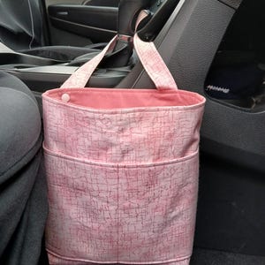 K&ouml;nnte beinhalten: Rosa Stoff-M&uuml;llbeutel f&uuml;r das Auto mit strukturiertem Muster und zwei Taschen. Die Tasche hat eine rosa Innenseite und h&auml;ngt am Schalthebel eines Autos.