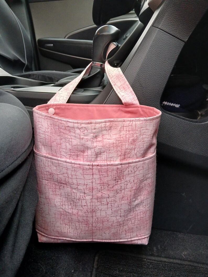 K&ouml;nnte beinhalten: Rosa Stoff-M&uuml;llbeutel f&uuml;r das Auto mit strukturiertem Muster und zwei Taschen. Die Tasche hat eine rosa Innenseite und h&auml;ngt am Schalthebel eines Autos.