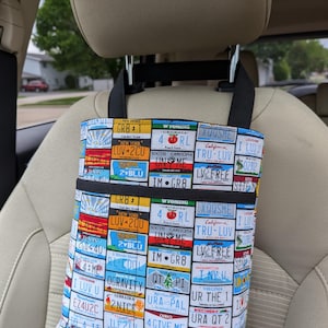 K&ouml;nnte beinhalten: Ein Autositz-Organizer mit einem farbenfrohen Nummernschildmuster. Der Organizer hat zwei schwarze Taschen und schwarze Tr&auml;ger zum Aufh&auml;ngen. Die Nummernschilder stammen aus verschiedenen Bundesstaaten, darunter New York, Wyoming und Louisiana.