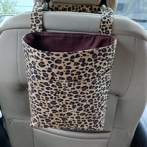 K&ouml;nnte beinhalten: Ein Autositz-Organizer mit Leopardenmuster. Der Organizer ist beige mit braunen Flecken und hat eine dunkelbraune Innenseite. Er wird an der Kopfst&uuml;tze befestigt und hat zwei Taschen.