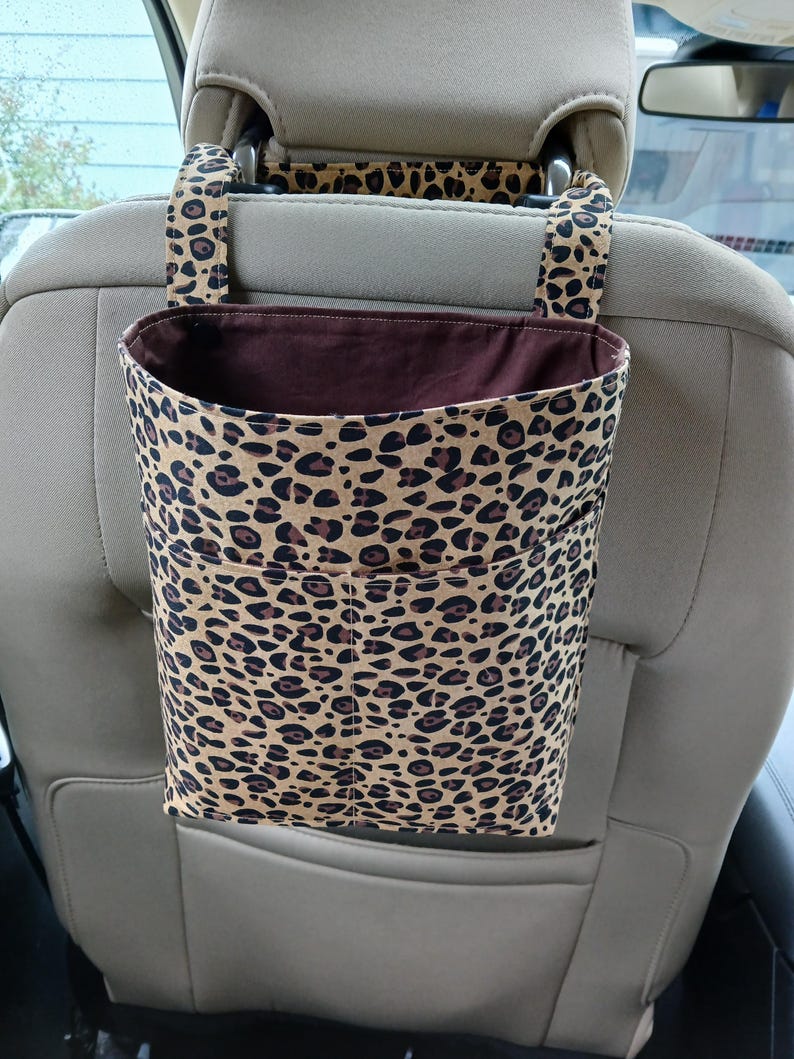 K&ouml;nnte beinhalten: Ein Autositz-Organizer mit Leopardenmuster. Der Organizer ist beige mit braunen Flecken und hat eine dunkelbraune Innenseite. Er wird an der Kopfst&uuml;tze befestigt und hat zwei Taschen.