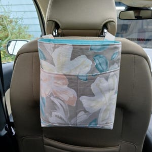 K&ouml;nnte beinhalten: Ein Autositz-Organizer mit einem Blumenmuster in Grau-, Pfirsich- und T&uuml;rkist&ouml;nen. Der Organizer hat eine hellblaue Innenseite und ist zum Aufh&auml;ngen an der Kopfst&uuml;tze konzipiert. Er verf&uuml;gt &uuml;ber eine Tasche auf der Vorderseite zur Aufbewahrung.