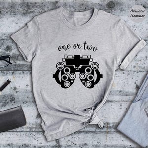 May include: A light gray short-sleeve t-shirt featuring a black graphic of an optometry phoropter and the text "one or two" in a cursive font. The shirt is labeled as Athletic Heather and is displayed with black glasses, a watch, a wallet, and jeans.
