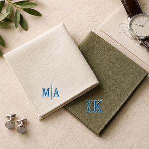 Puede incluir: Dos pañuelos de bolsillo plegados, uno color crema y otro verde oliva, cada uno con monogramas bordados en azul. El pañuelo crema muestra las letras M|A, mientras que el verde oliva muestra las letras VBK. Se incluyen dos gemelos plateados.