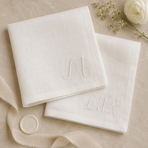 May include: Two square white fabric handkerchiefs featuring elegant white embroidery. One handkerchief displays the monogram JA, while the other features the monogram LWR. The fabric has a visible weave texture, and the edges are neatly finished.