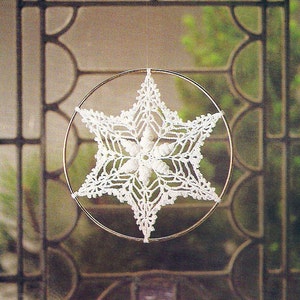 May include: A white crocheted snowflake hanging from a metal hoop. The snowflake is intricate and delicate, with a starburst design.