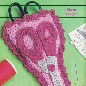May include: A pink and gray crocheted scissor holder with a scalloped edge. The holder is shaped like a pair of scissors and has a small flower detail at the top. The text "Shear Delight" is visible in the upper right corner of the image.