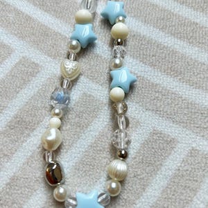 May include: A light blue and white beaded necklace with star and heart charms. The necklace is made with various sizes and shapes of beads, including clear, white, and silver beads.
