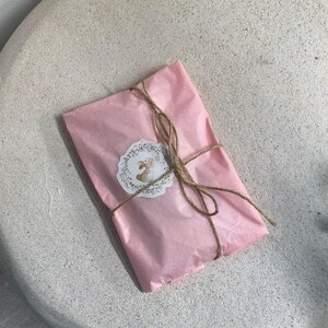 以下が含まれることがあります： ピンクのティッシュペーパーで包まれたパッケージで、紐で結ばれており、小さな丸いステッカーにはウサギのイラストが描かれています。