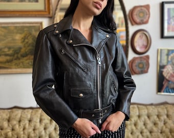 May include: A black leather motorcycle jacket with a belted waist and silver zippers. The jacket has a classic biker style with a notched collar and shoulder epaulets. The model is wearing a black and silver striped mini skirt.