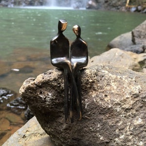 May include: A bronze sculpture of two figures sitting side-by-side on a rock. The figures are facing each other and appear to be in conversation. The sculpture is set against a backdrop of a waterfall and a lush green forest.
