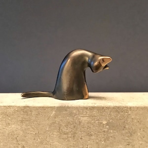 Puede incluir: Una pequeña escultura de bronce de un gato con la cabeza inclinada hacia un lado. El gato está sentado sobre una superficie de color marrón claro.