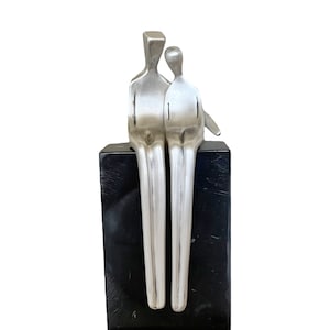 Puede incluir: Una escultura de plata de dos figuras estilizadas sentadas juntas sobre una base de mármol negro. Las figuras son abstractas y representan una pareja.