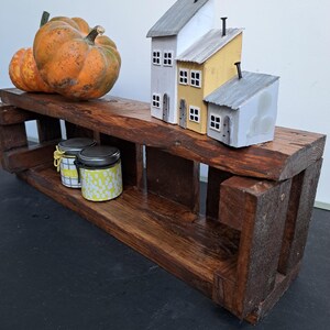 Pode incluir: Uma prateleira r&uacute;stica de madeira escura com objetos decorativos. Duas ab&oacute;boras em tons de laranja e amarelo est&atilde;o na prateleira superior. Tr&ecirc;s casas em miniatura com telhados cinza e amarelos tamb&eacute;m est&atilde;o na prateleira. Dois pequenos potes com tampas est&atilde;o na prateleira inferior.