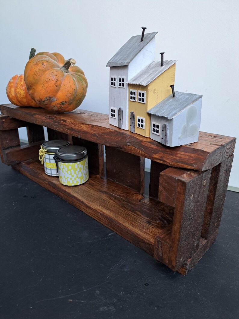 Pode incluir: Uma prateleira r&uacute;stica de madeira escura com objetos decorativos. Duas ab&oacute;boras em tons de laranja e amarelo est&atilde;o na prateleira superior. Tr&ecirc;s casas em miniatura com telhados cinza e amarelos tamb&eacute;m est&atilde;o na prateleira. Dois pequenos potes com tampas est&atilde;o na prateleira inferior.