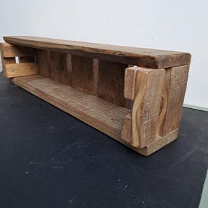 Pode incluir: Prateleira retangular de madeira com acabamento natural. A prateleira tem um compartimento longo e aberto e &eacute; constru&iacute;da com juntas vis&iacute;veis. O design apresenta uma borda superior ligeiramente curvada e suportes verticais.