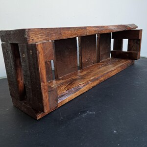 Pode incluir: Prateleira retangular de madeira r&uacute;stica com acabamento castanho escuro. A prateleira &eacute; constru&iacute;da com t&aacute;buas de madeira grossas, com um design de ripas nas laterais e na parte traseira. Projetada para montagem na parede ou coloca&ccedil;&atilde;o em uma superf&iacute;cie. Mede cerca de 40 cm de comprimento.