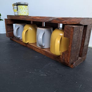 Pode incluir: Um suporte r&uacute;stico de madeira escura para canecas com quatro canecas de cer&acirc;mica. Duas canecas s&atilde;o amarelas e duas cinzentas. Um pequeno frasco com um padr&atilde;o amarelo e branco est&aacute; na prateleira superior.