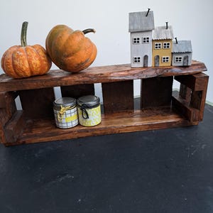 Pode incluir: Uma prateleira r&uacute;stica de madeira escura exibe duas ab&oacute;boras laranja, tr&ecirc;s casas em miniatura e dois pequenos potes. A prateleira est&aacute; sobre uma superf&iacute;cie preta. Os potes t&ecirc;m tampas decorativas.