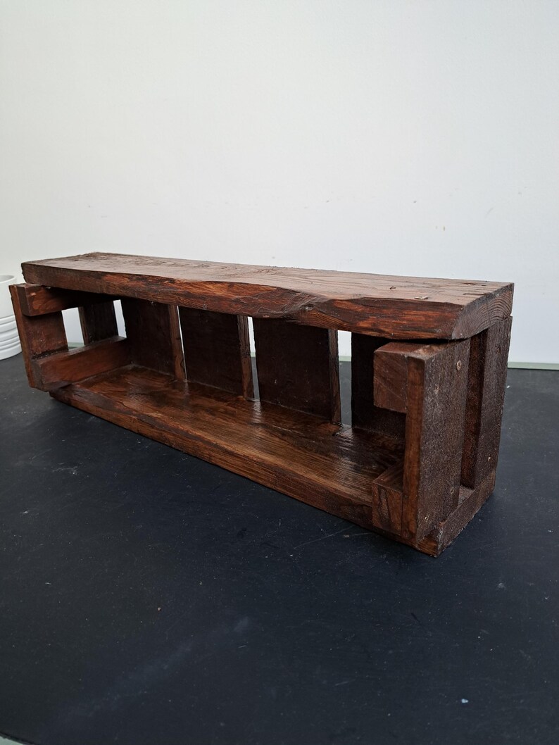 Pode incluir: Prateleira retangular de madeira castanha escura com borda natural na parte superior. A prateleira tem suportes verticais que criam compartimentos abertos. A madeira tem um tom rico e quente e gr&atilde;o vis&iacute;vel, sugerindo um design artesanal.