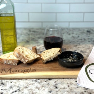 May include: A wooden serving board with the word "Mangia" engraved on it. The board has three slices of bread, a small bowl of olive oil, and a glass of red wine. A white towel with the word "Olive" printed on it is in the background.