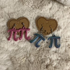 May include: Two pairs of dangle earrings featuring the mathematical symbol pi. One pair is pink with glitter, the other is blue with a gradient effect. Each earring hangs from a small, heart-shaped cardboard backing.