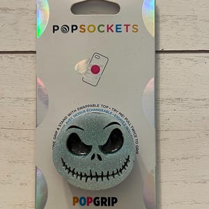 May include: A PopSockets PopGrip featuring a Jack Skellington design. The grip is light blue with glitter and a black face. The packaging includes the PopSockets logo and text that reads "One grip & stand with swappable top".