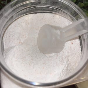 K&ouml;nnte beinhalten: Ein durchsichtiger Plastikbeh&auml;lter gef&uuml;llt mit wei&szlig;em Pulver und einer wei&szlig;en Plastikschaufel. Das Pulver hat einen leicht rosafarbenen Farbton.