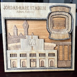 May include: A wooden model of Jordan-Hare Stadium in Auburn, Alabama. The model features a detailed depiction of the stadium, including the seating, the field, and the surrounding buildings. The model is painted in a variety of shades of brown and beige.