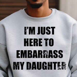 Puede incluir: Sudadera gris claro con el texto negro "I'M JUST HERE TO EMBARRASS MY DAUGHTER". El texto está en letras negras. La sudadera tiene cuello redondo y mangas largas.