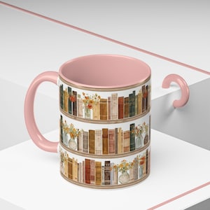 May include: A ceramic mug with a pink interior and handle. The exterior features a watercolor design of bookshelves filled with books and vases of flowers. The mug is a light pink color.