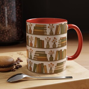May include: A ceramic mug with a red interior and handle, featuring a repeating pattern of bookshelves with books and vases of flowers. The books are in shades of brown and green, and the flowers are orange.