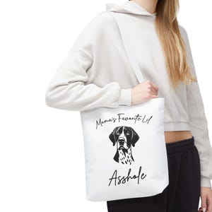 May include: White tote bag with a black and white dog graphic and the text "Mama's Favorite Lif" and "Asshole". The bag has a shoulder strap and is being held by a person.