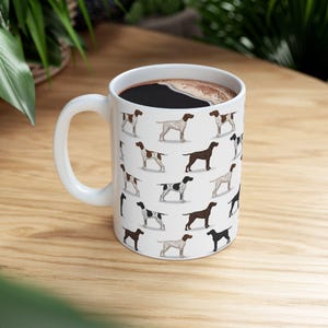 May include: White ceramic coffee mug featuring a repeating pattern of various dog breeds in brown, black, and white. The mug is filled with dark coffee and has a white handle. The mug is sitting on a wooden surface.