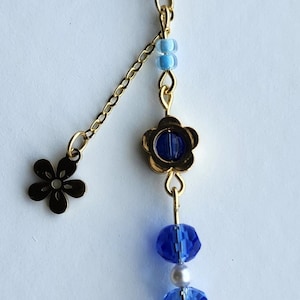 May include: A gold key chain with a blue and white beaded charm. The charm features a flower with a blue center, blue crystal beads, and a white flower with blue accents. The key chain also has a small gold flower charm.