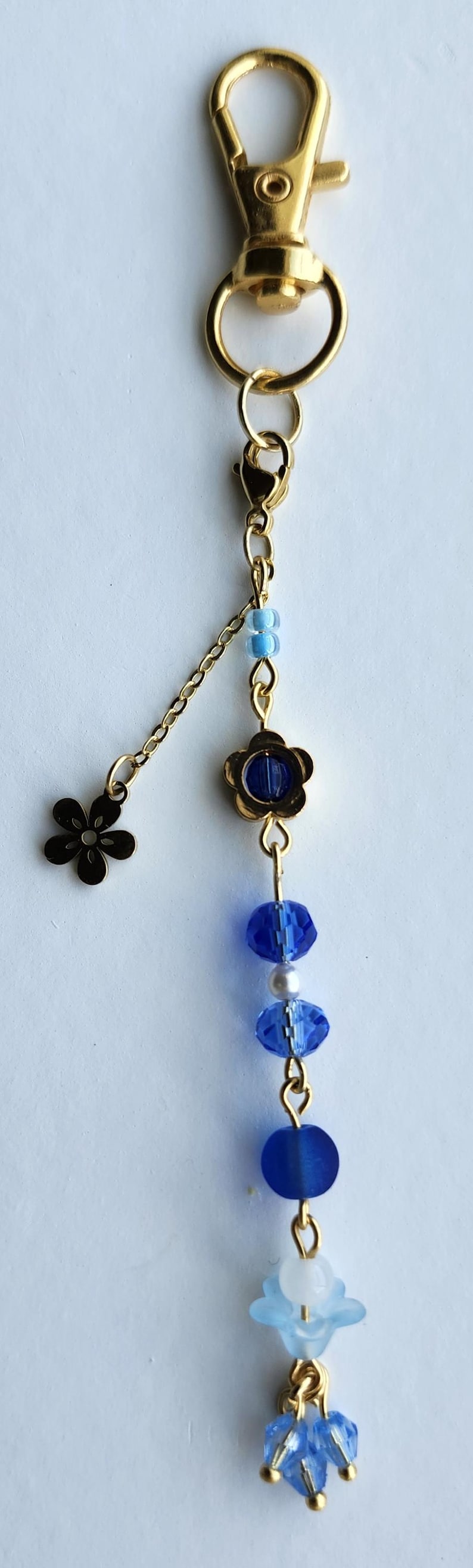 May include: A gold key chain with a blue and white beaded charm. The charm features a flower with a blue center, blue crystal beads, and a white flower with blue accents. The key chain also has a small gold flower charm.