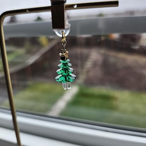 Op de afbeelding: Een kerstboomoorbel met een goudkleurige ster en een groen, facetgeslepen boomontwerp. De oorbel hangt aan een goudkleurige haak. De oorbel wordt tentoongesteld op een goudkleurige standaard.