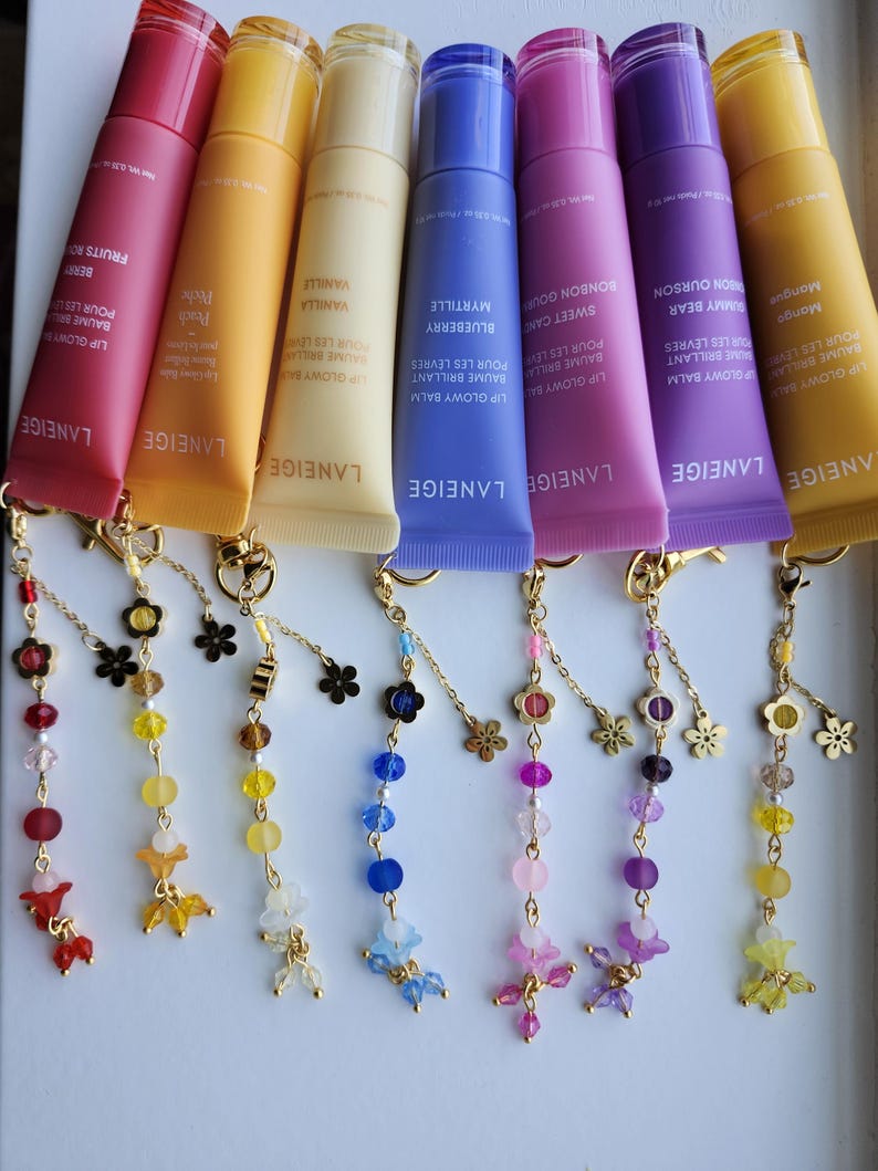 May include: Eight colorful lip balm tubes with gold charms attached. The lip balms are labeled with different flavors and colors, including mango, honey bear, sweet candy, blueberry, vanilla, peach, and berry. The charms are made of gold chains and beads with small flower accents.