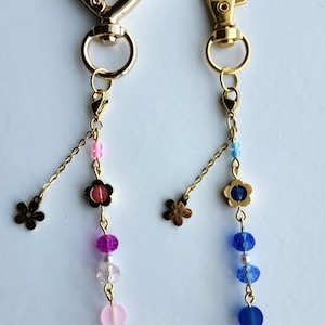 May include: Two gold keychains with different clasps. The keychain on the left has a heart clasp and a pink and purple beaded charm. The keychain on the right has a fish hook clasp and a blue and white beaded charm. Both charms have a small flower charm at the top.